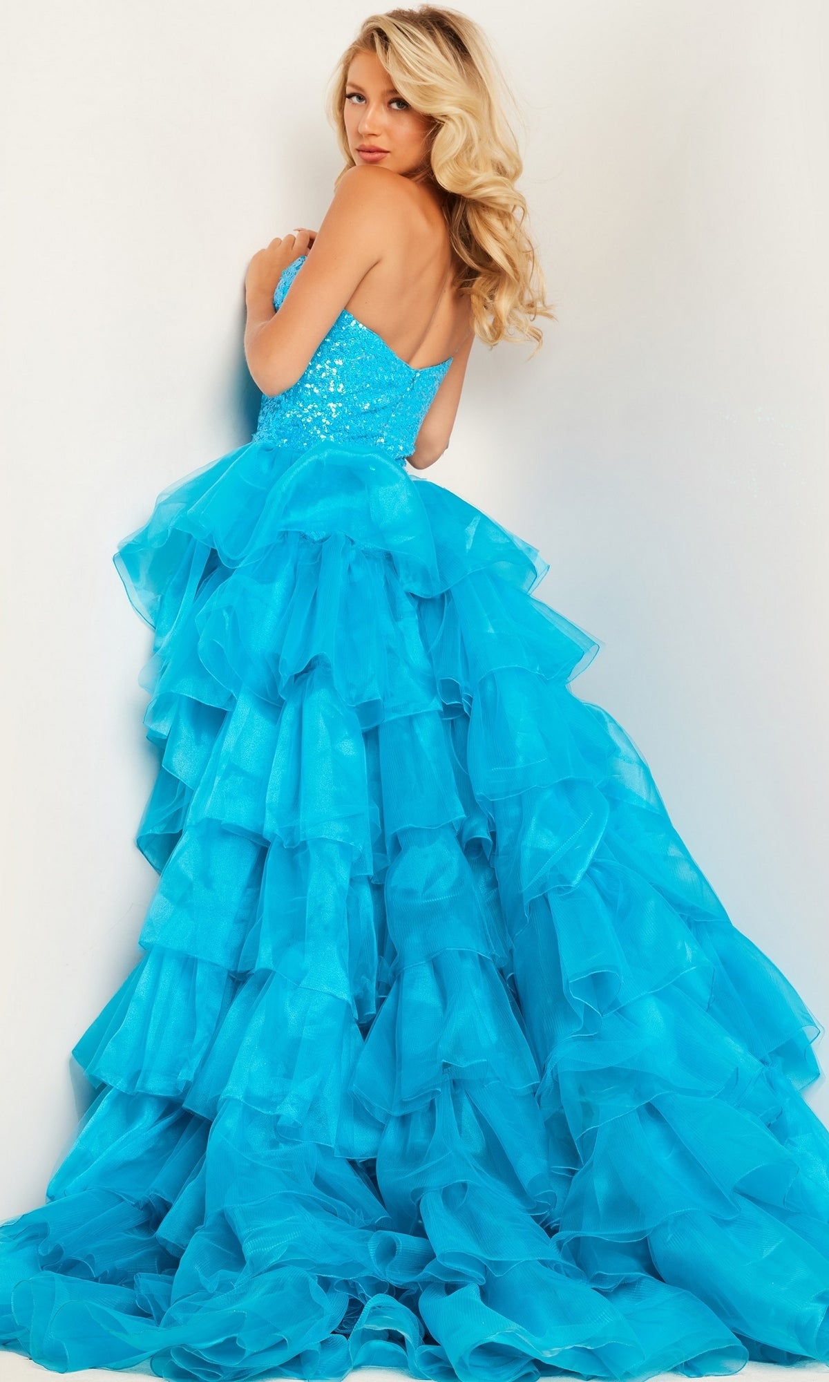 A woman poses against a light background, looking over her shoulder in the Jovani 37689 Ruffled Long Sequin Pageant Dress, featuring a strapless, bright blue design with sequins and ruffled accents.